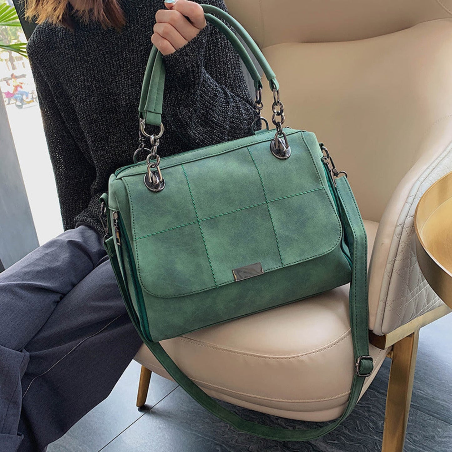 Woman holding green matte PU leather large capacity Boston shoulder tote bag by top handles with silver chain hardware and quilted stitch detail, resting on a beige chair — Avenue
