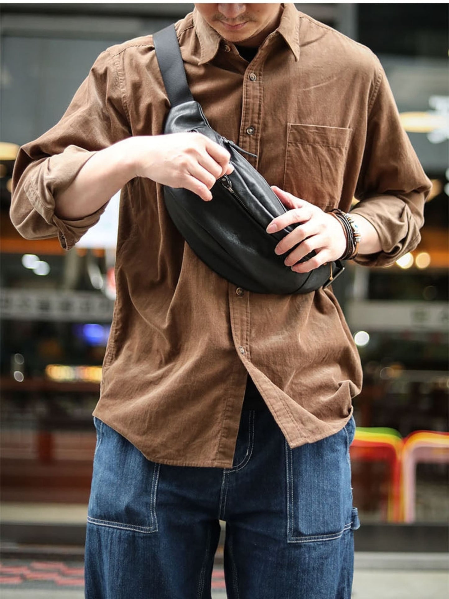 Man wearing black plant-tanned cow leather crossbody mobile phone bag over shoulder opening zipper in urban street setting — Avenue