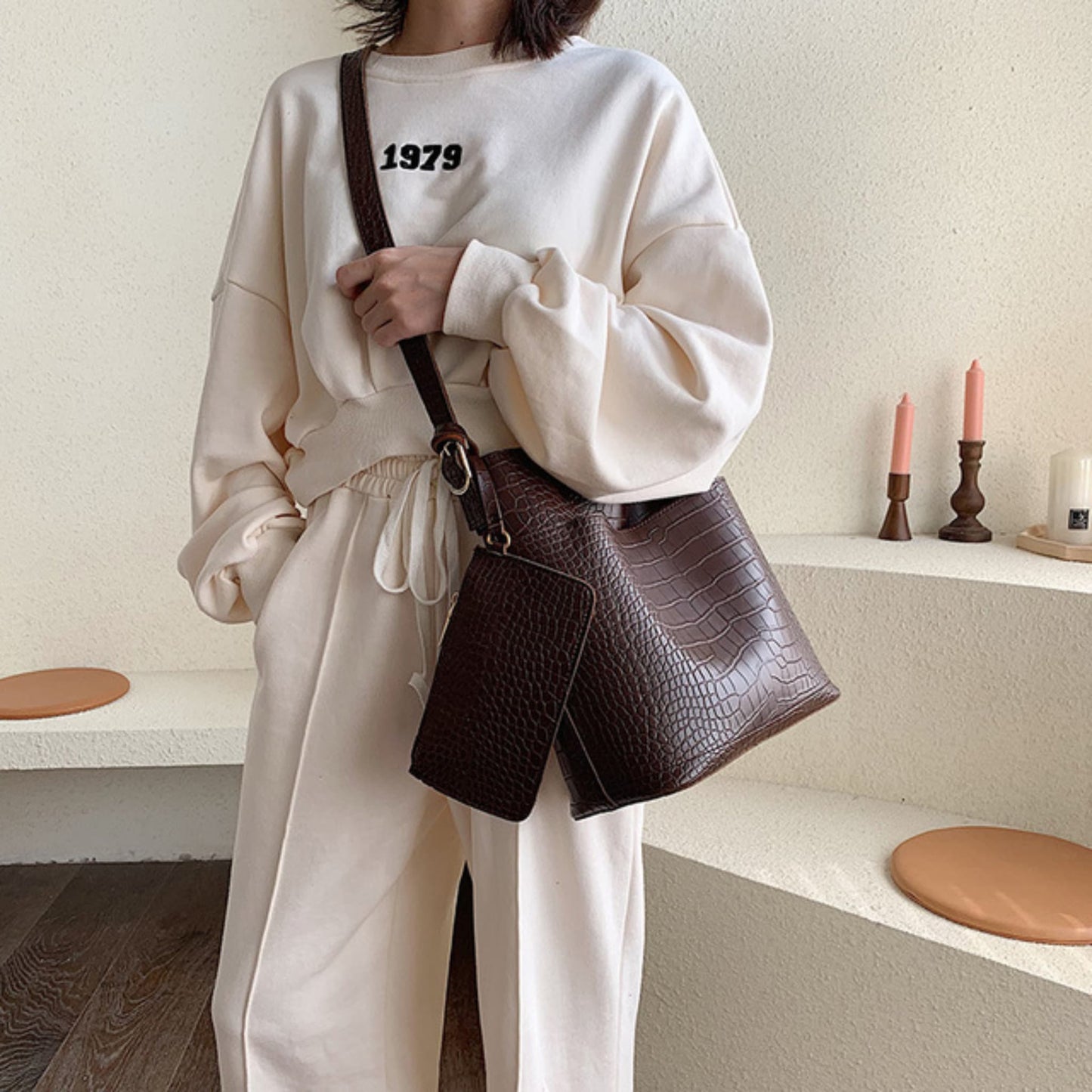 Woman wearing a women's one shoulder bucket bag in coffee, croc-embossed PU leather with magnetic snap closure, styled with a cream oversized sweatsuit | Avenue