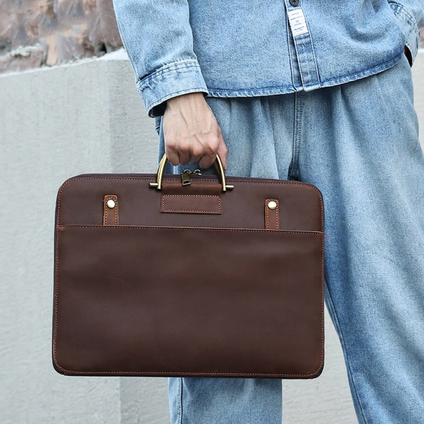 Avenue Crazy Horse Genuine Leather Briefcase for Men – Lifestyle shot in 2121 Brown, held by a man in denim outfit, showcasing antique brass top handle grip, brass stud detailing, front slip pocket, and contrast stitching against an outdoor stone background