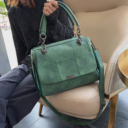 Woman holding green matte PU leather large capacity Boston shoulder tote bag by top handles with silver chain hardware and quilted stitch detail, resting on a beige chair — Avenue