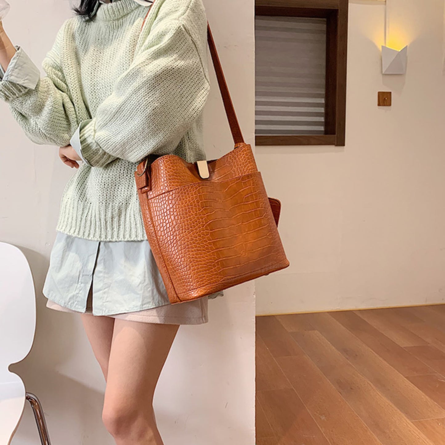 Woman wearing a women's one shoulder bucket bag in brown, croc-embossed PU leather with magnetic snap closure, styled with a green knit sweater and denim skirt | Avenue