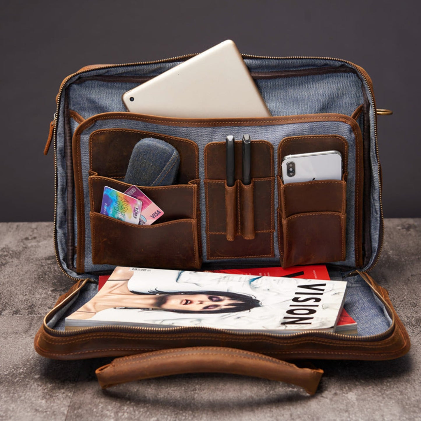Avenue Men's Large Capacity Crazy Horse Leather Laptop Briefcase in dark brown — interior organisation view showing blue fabric lining, tablet pocket at top, card slots and accessory pocket on left panel, dual pen holders and smartphone pocket on right panel, magazine and documents in main compartment, brass zipper pulls on both sides, and leather top handle at bottom