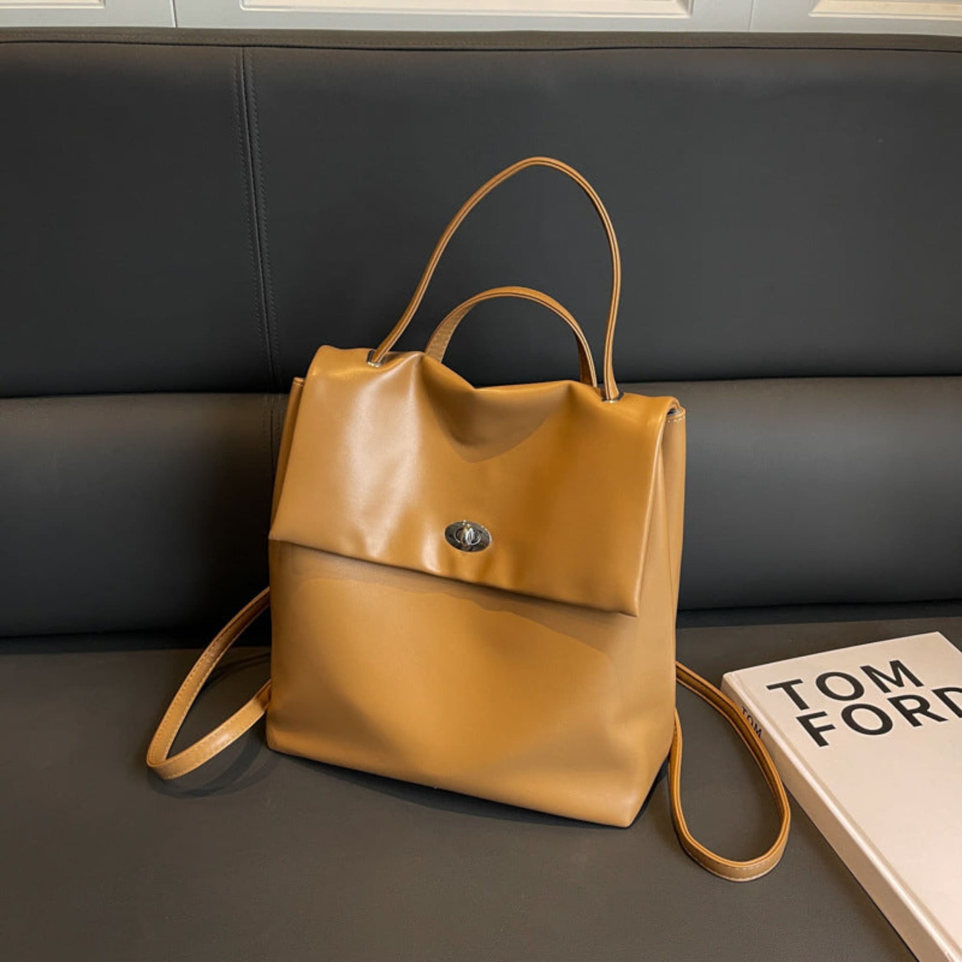 Women's Retro Flap Backpack in Brown – Lifestyle Shot on Sofa Styled with Tom Ford Book, Turn-Lock Closure and Dual Backpack Straps | Avenue