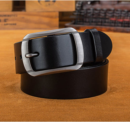 Men's full-grain cowhide leather belt in black with silver pin buckle, coiled and laying flat on tan leather surface with wooden background | Avenue