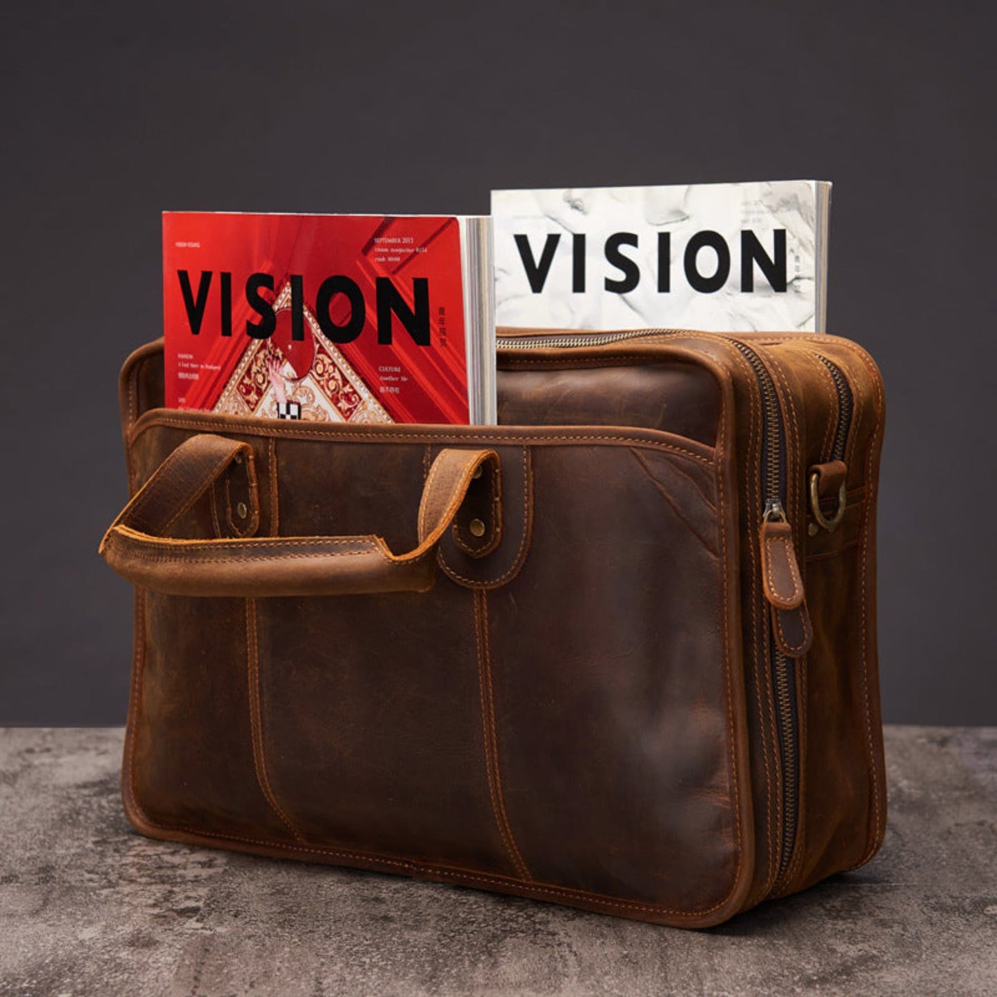 Avenue Men's Large Capacity Crazy Horse Leather Laptop Briefcase in dark brown — top-open view demonstrating large capacity with two magazines stored inside, showing main zipper opening, front flap pocket with brass stud closure, main zipper pull visible on right side, shoulder strap D-ring attachment, and contrast topstitching, styled on concrete surface against dark grey background