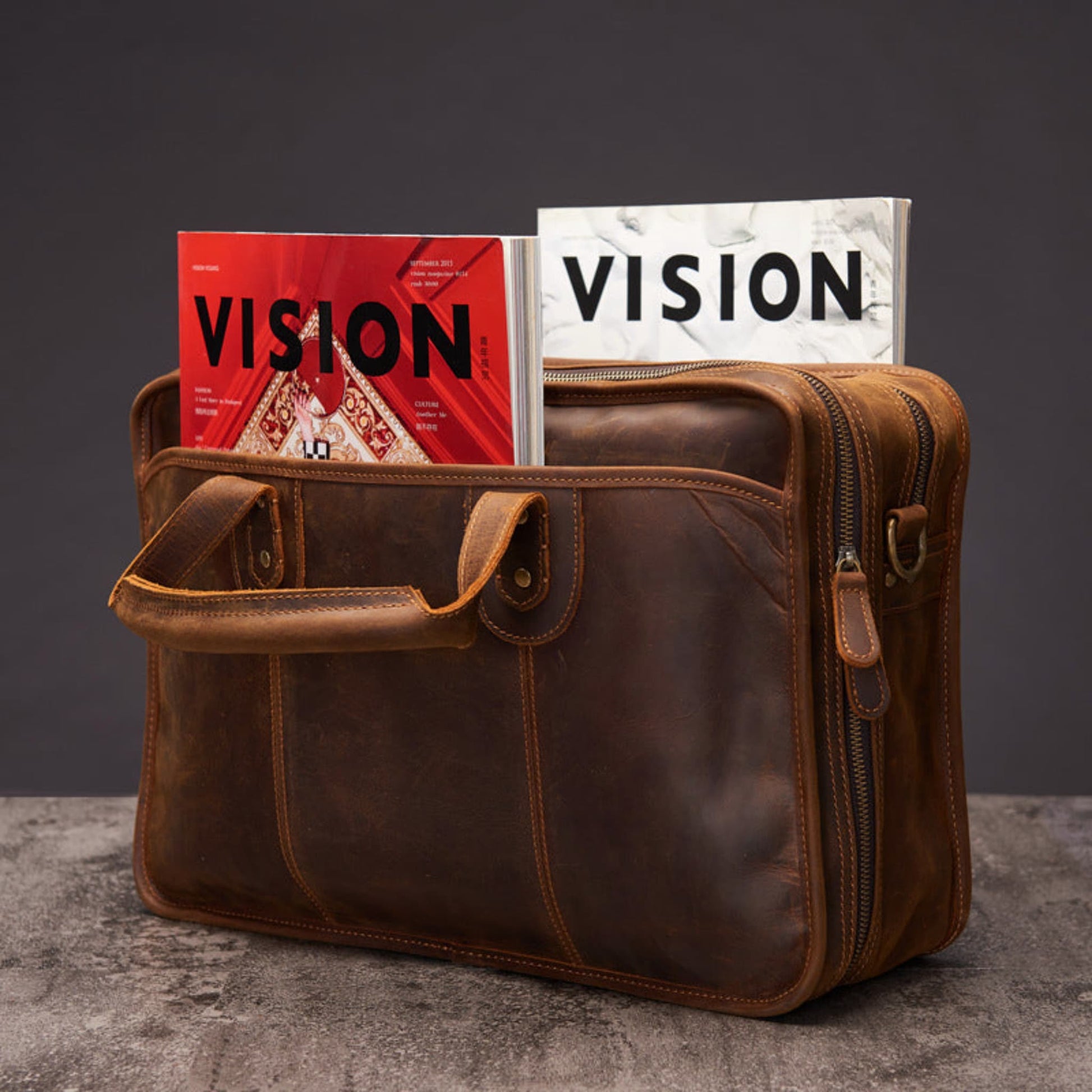 Avenue Men's Large Capacity Crazy Horse Leather Laptop Briefcase in dark brown — top-open view demonstrating large capacity with two magazines stored inside, showing main zipper opening, front flap pocket with brass stud closure, main zipper pull visible on right side, shoulder strap D-ring attachment, and contrast topstitching, styled on concrete surface against dark grey background