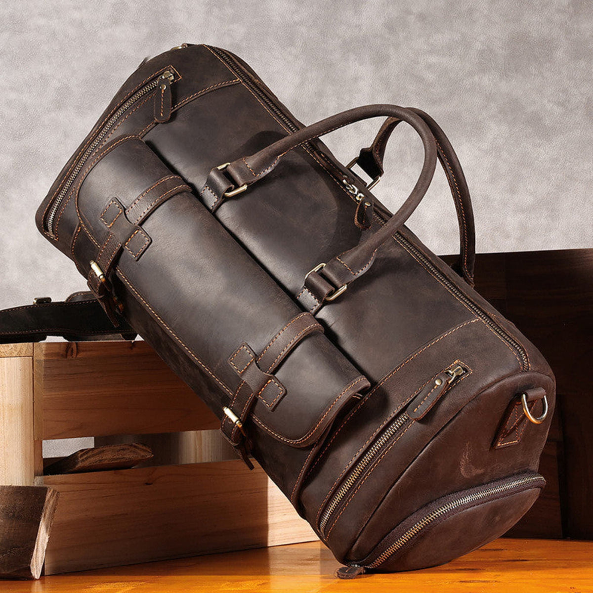 Avenue’s coffee Top-Grain Cowhide Leather Travel Duffle Bag with twin carry handles, displayed on wooden furniture against a textured grey background.