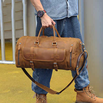 Man carrying Avenue’s brown Top-Grain Cowhide Leather Travel Duffle Bag by its twin handles outdoors against a modern building background.