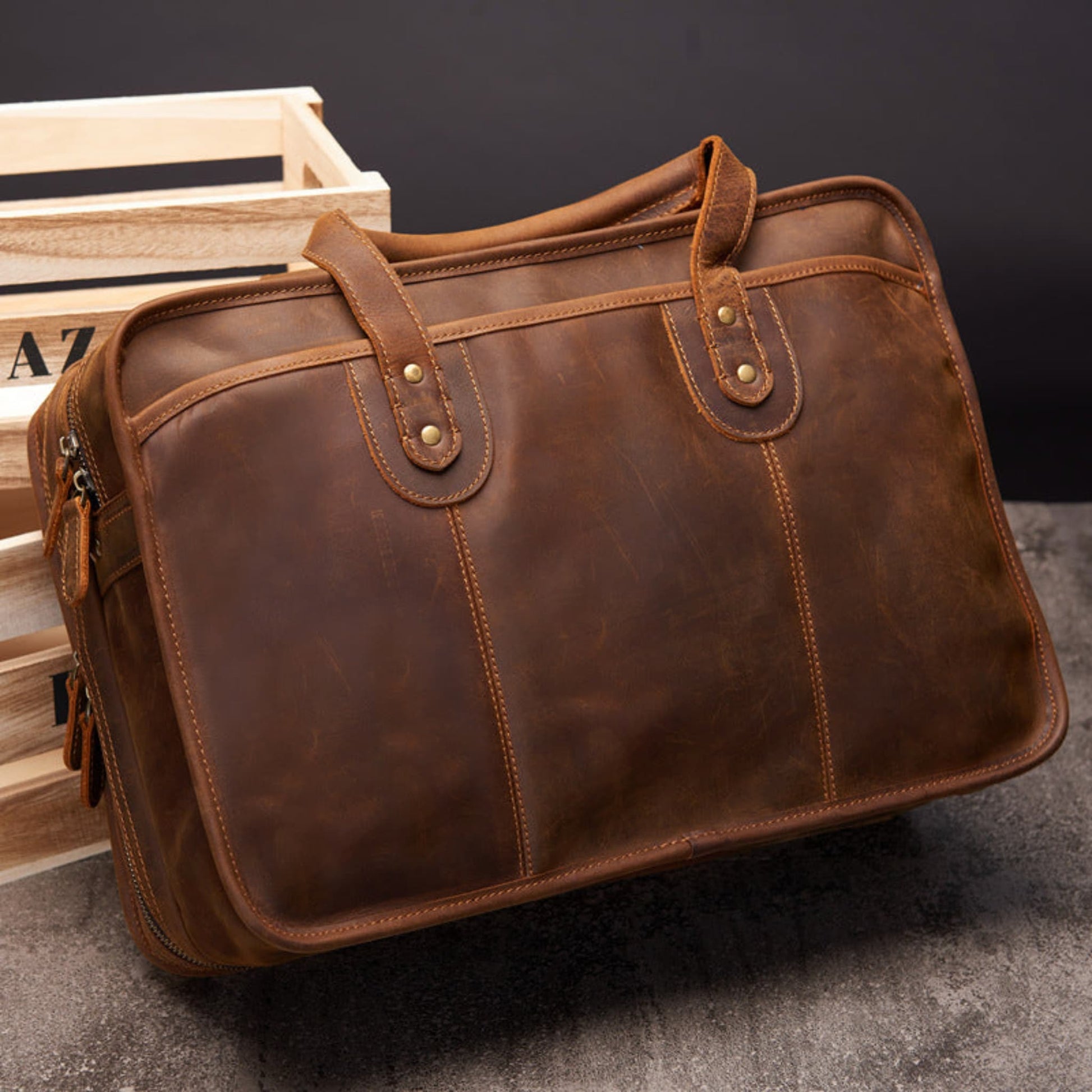 Avenue Men's Large Capacity Crazy Horse Leather Laptop Briefcase in dark brown — angled front view showcasing natural crazy horse leather patina, brass stud hardware on dual handle loops, contrast topstitching, zipper pulls on left side, and large structured body, styled against rustic wooden crate on dark background