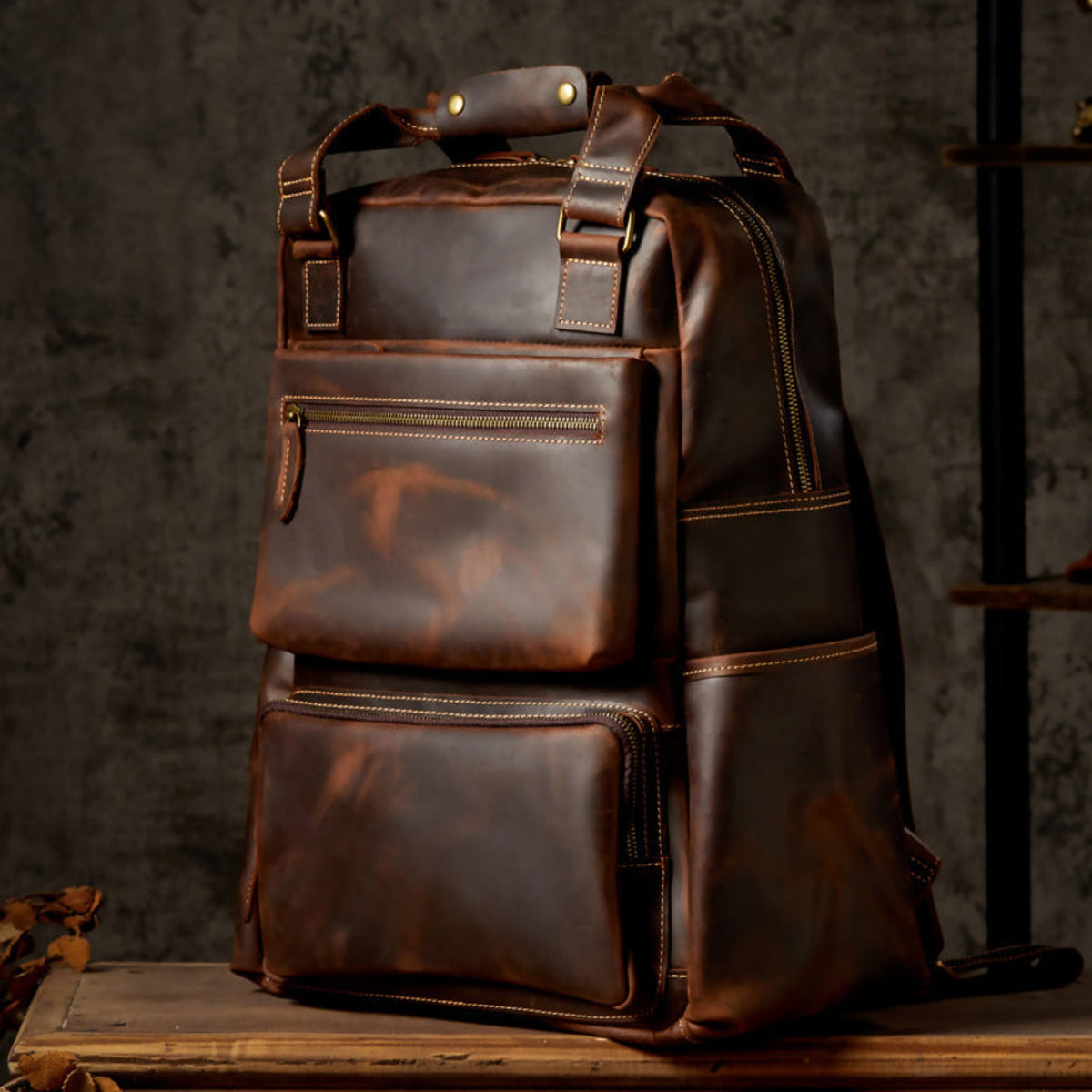 Unisex handmade retro crazy horse leather oversized backpack in brown, three-quarter view showing leather top handle,antique brass rivets and rich patina on wooden surface | Avenue
