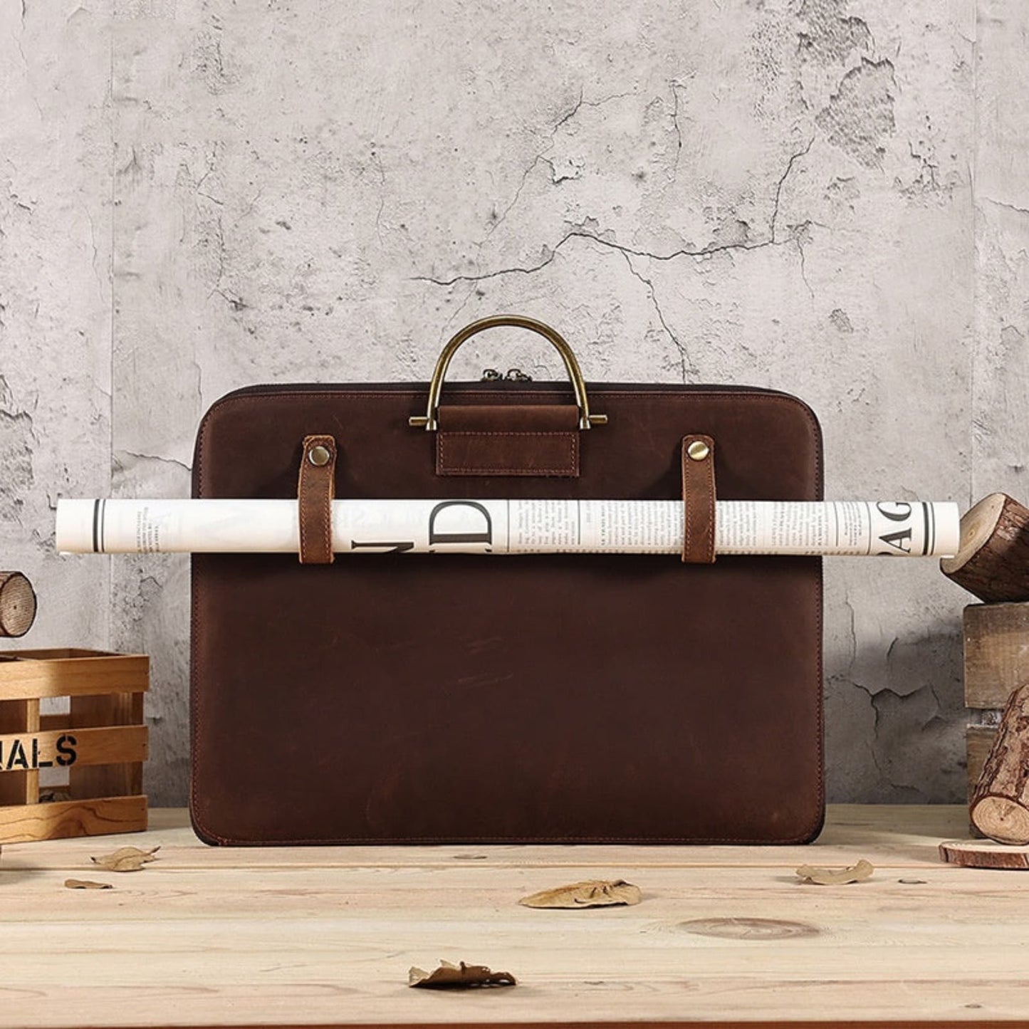 Avenue Crazy Horse Genuine Leather Briefcase for Men – Front view in 2121 Brown showcasing front leather strap holding a rolled newspaper, antique brass top handle and stud detailing, styled on a wooden surface against a rustic stone wall with wooden props